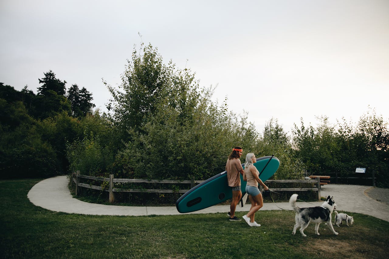 A couple walking their dogs while carrying a paddle board through a lush park during the day.